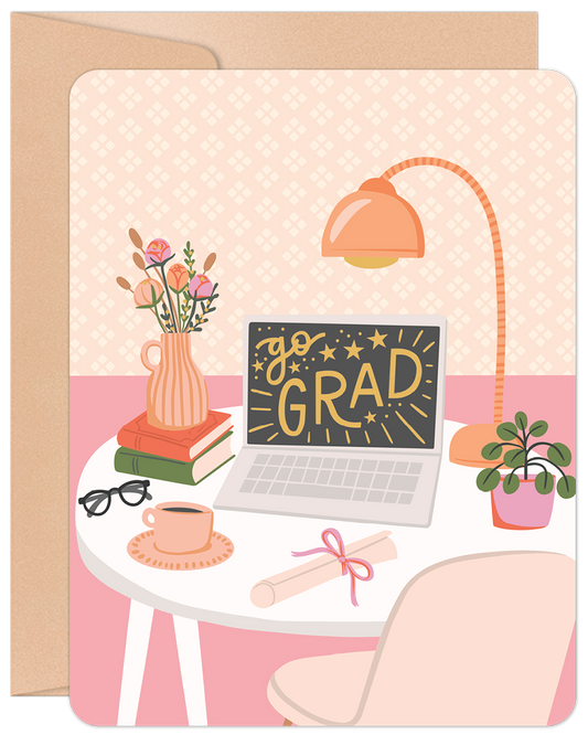 Front of a graduation greeting card showing a desk with a laptop displaying 'Go Grad!', books, flowers, and a diploma.