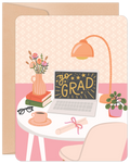 Close-up of an encouraging congratulations card featuring a stylish desk setup with a laptop displaying 'go GRAD' surrounded by stars, a stack of books, a pink vase with flowers, a task lamp, a coffee cup, glasses, and a rolled diploma scroll, all on a pastel pink background.
