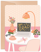 Close-up of an encouraging congratulations card featuring a stylish desk setup with a laptop displaying 'go GRAD' surrounded by stars, a stack of books, a pink vase with flowers, a task lamp, a coffee cup, glasses, and a rolled diploma scroll, all on a pastel pink background.