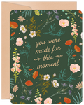 Front of an inspiring graduation greeting card featuring a dark green background with a vibrant botanical border of orange poppies, pink bellflowers, white blossoms, and green foliage. The central text reads 'you were made for this moment' in elegant script, accompanied by a kraft paper envelope.