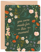 Front of an inspiring graduation greeting card featuring a dark green background with a vibrant botanical border of orange poppies, pink bellflowers, white blossoms, and green foliage. The central text reads 'you were made for this moment' in elegant script, accompanied by a kraft paper envelope.