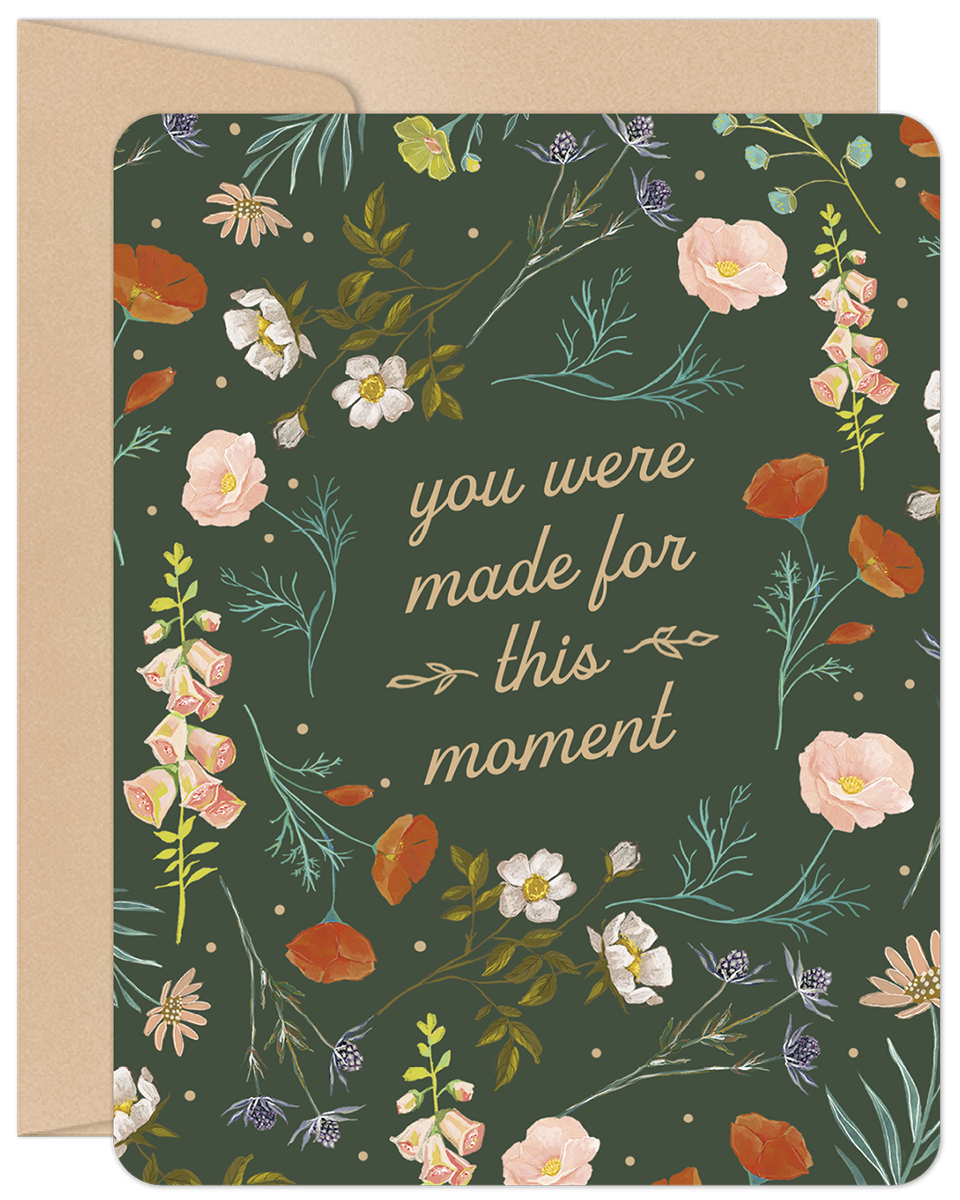 Front of an inspiring graduation greeting card featuring a dark green background with a vibrant botanical border of orange poppies, pink bellflowers, white blossoms, and green foliage. The central text reads 'you were made for this moment' in elegant script, accompanied by a kraft paper envelope.
