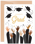 Front of a modern graduation greeting card featuring 'Congrats Grad' in gold typography, surrounded by black tossed graduation caps, gold stars, and diverse hands raised in celebration, set against a white background with a kraft envelope visible.