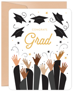 Front of a modern graduation greeting card featuring 'Congrats Grad' in gold typography, surrounded by black tossed graduation caps, gold stars, and diverse hands raised in celebration, set against a white background with a kraft envelope visible.