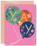 Front of a vibrant pink greeting card featuring three floral-patterned balloons spelling 'YAY' in gold letters, with a kraft brown envelope peeking out.