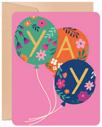 Front of a vibrant pink greeting card featuring three floral-patterned balloons spelling 'YAY' in gold letters, with a kraft brown envelope peeking out.