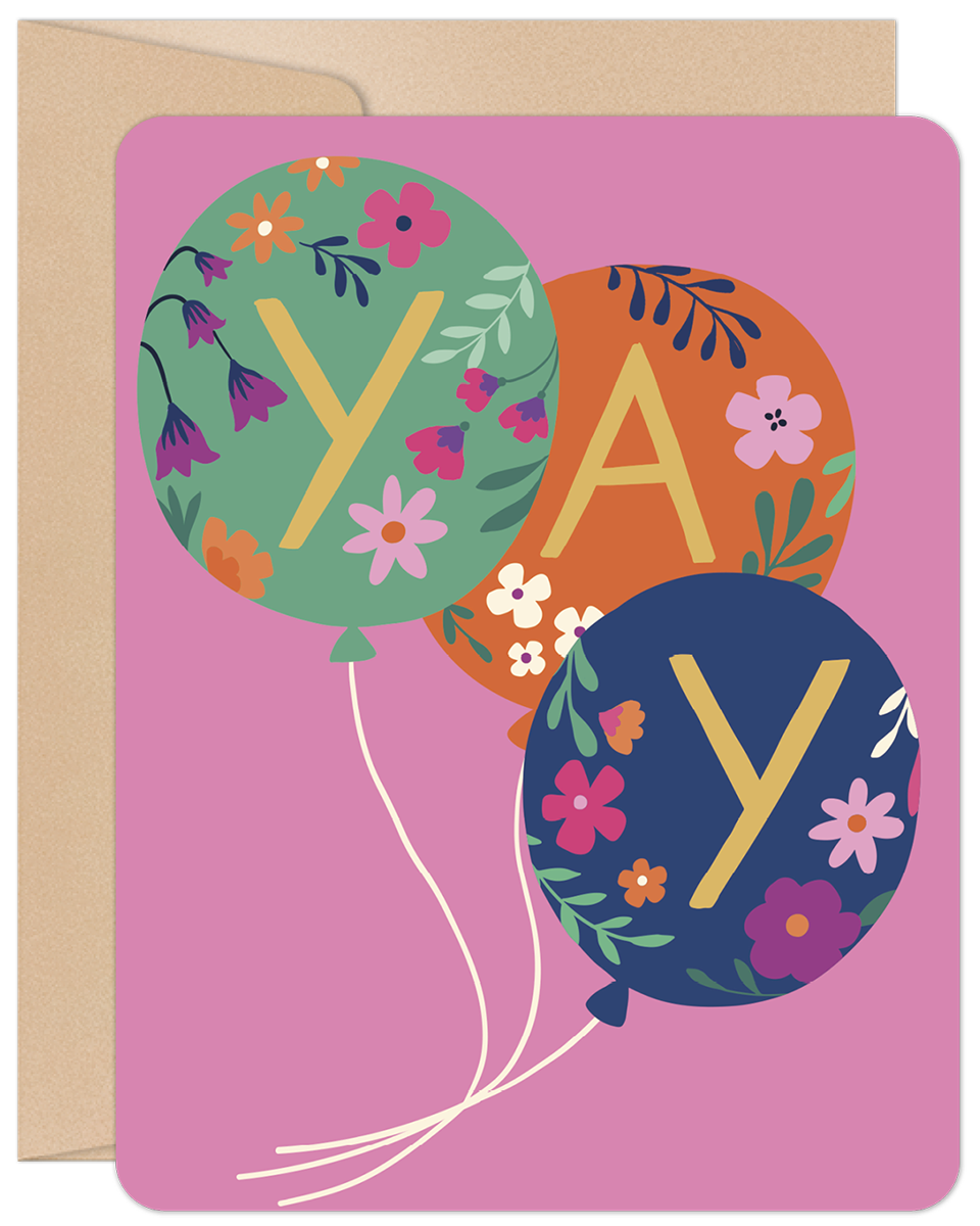 Front of a vibrant pink greeting card featuring three floral-patterned balloons spelling 'YAY' in gold letters, with a kraft brown envelope peeking out.