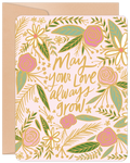 Elegant anniversary greeting card featuring a pink background, metallic gold 'May your love always grow' script, and vibrant floral illustrations of pink roses, white daisies, and green leaves, paired with a natural kraft envelope.