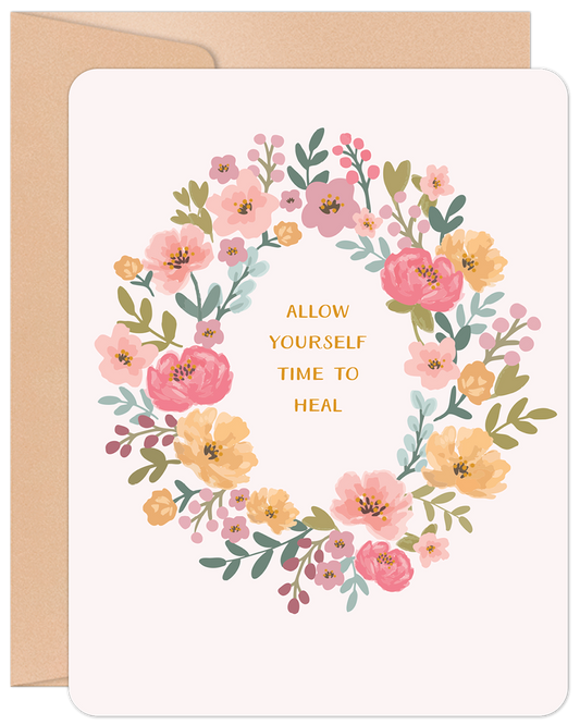 Floral wreath greeting card with pink and yellow flowers surrounding 'ALLOW YOURSELF TIME TO HEAL' message, on a kraft envelope.