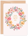 Front of a gentle encouragement greeting card featuring a watercolor floral wreath in pink, yellow, and green, with the heartfelt message 'ALLOW YOURSELF TIME TO HEAL' in the center, resting on a light cream background with a craft paper envelope visible behind.