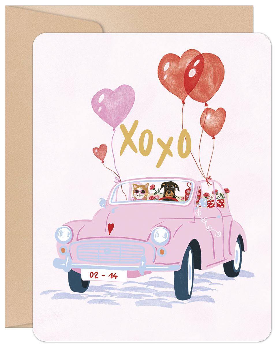 Front of a Valentine's Day greeting card featuring a charming illustration of a cat driving a pink vintage car with a dog in the passenger seat. The car is adorned with red and pink heart-shaped balloons, "XOXO" text, and a "02-14" license plate, all on a light pink background with a kraft paper envelope behind it.