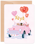Front of a Valentine's Day greeting card featuring a charming illustration of a cat driving a pink vintage car with a dog in the passenger seat. The car is adorned with red and pink heart-shaped balloons, "XOXO" text, and a "02-14" license plate, all on a light pink background with a kraft paper envelope behind it.