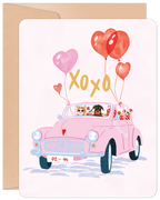 Front of a Valentine's Day greeting card featuring a charming illustration of a cat driving a pink vintage car with a dog in the passenger seat. The car is adorned with red and pink heart-shaped balloons, "XOXO" text, and a "02-14" license plate, all on a light pink background with a kraft paper envelope behind it.