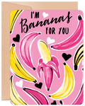 Front of a vibrant pink greeting card featuring playful illustrations of yellow and pink bananas, accented with black and white hearts. Bold handwritten text reads "I'M Bananas FOR YOU". The card is slightly angled, revealing a corner of a brown kraft envelope.