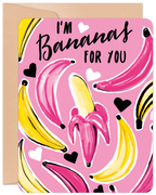 Front of a vibrant pink greeting card featuring playful illustrations of yellow and pink bananas, accented with black and white hearts. Bold handwritten text reads "I'M Bananas FOR YOU". The card is slightly angled, revealing a corner of a brown kraft envelope.