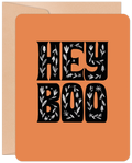 Front of a vibrant orange greeting card for Halloween, featuring decorative black text that reads 'HEY BOO' with intricate white floral and leaf patterns. A brown kraft envelope is partially visible. Ideal for sending cute spooky season wishes to friends.
