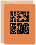 Front of a vibrant orange greeting card for Halloween, featuring decorative black text that reads 'HEY BOO' with intricate white floral and leaf patterns. A brown kraft envelope is partially visible. Ideal for sending cute spooky season wishes to friends.