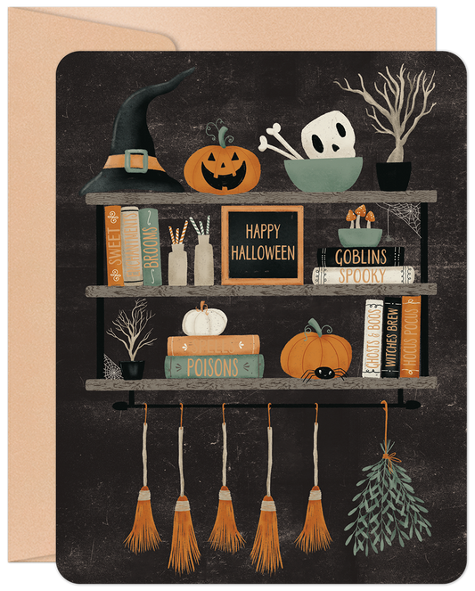 Front of a charming Halloween greeting card featuring a witch's hat, carved pumpkins, a skull, and magic books on rustic shelves.