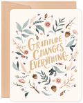 Front of a heartfelt Thanksgiving greeting card with 'Gratitude Changes Everything' in elegant calligraphy, surrounded by an autumn wreath of botanical leaves, acorns, and berries, presented with a neutral kraft envelope.