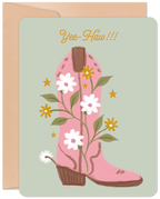 Front of an encouraging birthday greeting card featuring a pink cowboy boot adorned with white and yellow daisies, green vines, and gold stars on a light green background, with 'Yee-Haw!!!' in orange script. A kraft paper envelope is visible behind the card.