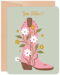 Front of an encouraging birthday greeting card featuring a pink cowboy boot adorned with white and yellow daisies, green vines, and gold stars on a light green background, with 'Yee-Haw!!!' in orange script. A kraft paper envelope is visible behind the card.
