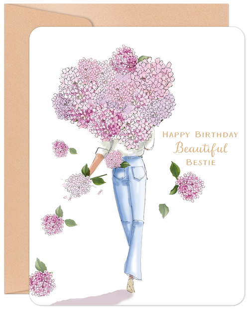 Front of a charming birthday greeting card featuring an illustration of a woman in jeans, her head replaced by a voluminous pink hydrangea bouquet, with scattered petals and the golden text 'Happy Birthday Beautiful Bestie' against a white background, accompanied by a kraft envelope.