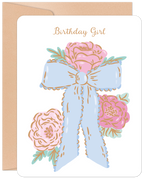 Elegant greeting card for a girl with 'Birthday Girl' in gold script, featuring a large pastel blue bow and soft pink floral bouquet on a white background, accompanied by a kraft envelope.