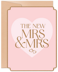 Front of a modern pink wedding greeting card for two brides, featuring gold glitter text 'THE NEW MRS & MRS' within a large white heart, bordered by a subtle gold trim, with a kraft envelope visible behind.