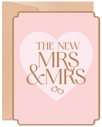 Front of a modern pink wedding greeting card for two brides, featuring gold glitter text 'THE NEW MRS & MRS' within a large white heart, bordered by a subtle gold trim, with a kraft envelope visible behind.