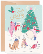 Whimsical Christmas greeting card featuring five cartoon dogs playing in the snow, building a snowman, and admiring a decorated Christmas tree with a star. Holly and 'Tis the Season' text are visible. Card has rounded corners and a brown envelope.