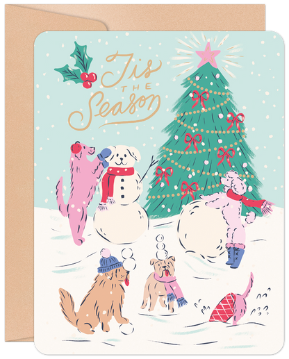 Front of a 'Tis The Season Christmas greeting card featuring cute dogs building a snowman and playing in the snow by a decorated Christmas tree.