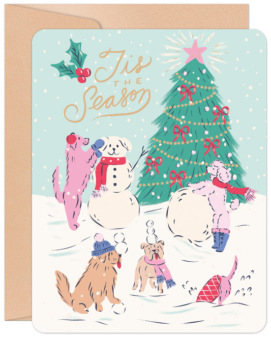 Front of a 'Tis The Season Christmas greeting card featuring cute dogs building a snowman and playing in the snow by a decorated Christmas tree.