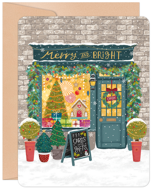 Front of a festive holiday greeting card featuring a charming storefront decorated with Christmas garlands, a wreath, sparkling lights, a gingerbread house, and a snowy sidewalk. The shop window displays a decorated tree and gifts with text 'Merry and Bright'.