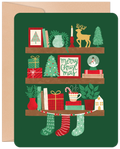 Front of a festive green holiday greeting card featuring three decorated shelves with charming illustrations of gifts, mini Christmas trees, a reindeer, snowman globe, candles, mugs, and stockings, perfect for spreading cheer.