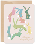 A cute greeting card with a light pink background featuring multiple stylized pastel bunnies in various shades of pink, blue, green, and yellow, each with unique decorative patterns and fluffy white tails. The bottom right corner has gold script text that reads 'There's no bunny like you!'. A brown kraft envelope is partially visible on the left side. Ideal for sending cheerful spring or celebration wishes.