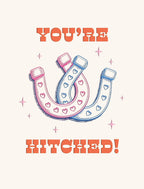 Front of a cute wedding greeting card with 'YOU'RE HITCHED!' in bold orange text, featuring two interlocking pink and blue horseshoes adorned with small hearts and surrounded by tiny sparkling stars.