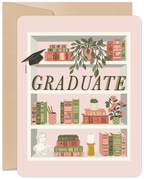 Front of a graduation greeting card featuring a charming bookshelf illustration. The shelves are filled with classic books, potted plants, a classical bust, a candle, and a prominent graduation cap. The word "GRADUATE" is displayed in large, elegant letters in the center.