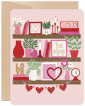 Front of a romantic greeting card with a bookshelf design, featuring three shelves adorned with pink and red Valentine's decorations like hearts, 'XOXO', 'Be Mine' signs, flowers, books, and plants, all on a light pink background.