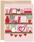 Front of a romantic greeting card with a bookshelf design, featuring three shelves adorned with pink and red Valentine's decorations like hearts, 'XOXO', 'Be Mine' signs, flowers, books, and plants, all on a light pink background.