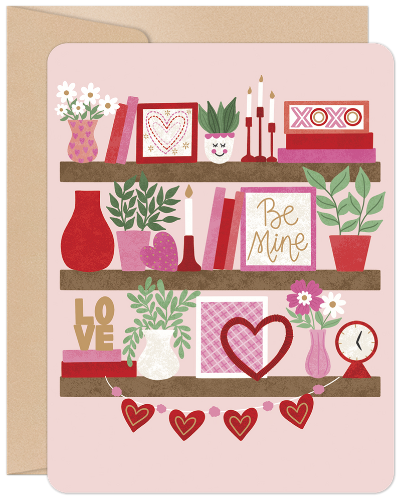 Front of a romantic greeting card with a bookshelf design, featuring three shelves adorned with pink and red Valentine's decorations like hearts, 'XOXO', 'Be Mine' signs, flowers, books, and plants, all on a light pink background.