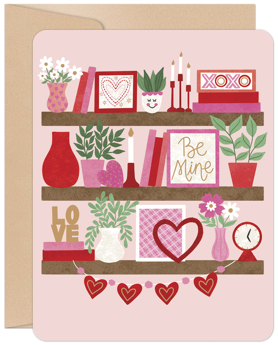 Front of a romantic greeting card with a bookshelf design, featuring three shelves adorned with pink and red Valentine's decorations like hearts, 'XOXO', 'Be Mine' signs, flowers, books, and plants, all on a light pink background.