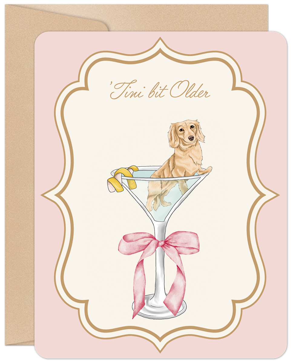Front of a funny birthday greeting card featuring a cute golden dachshund puppy sitting in a martini glass with a lemon twist, on a light pink background with an ornate gold frame. Text reads "'Tini bit Older".