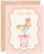 Front of a funny birthday greeting card featuring a cute golden dachshund puppy sitting in a martini glass with a lemon twist, on a light pink background with an ornate gold frame. Text reads "'Tini bit Older".