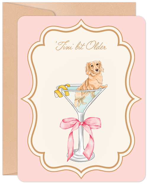 Front of a funny birthday greeting card featuring a cute golden dachshund puppy sitting in a martini glass with a lemon twist, on a light pink background with an ornate gold frame. Text reads "'Tini bit Older".