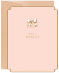A modern blush pink greeting card with a detailed gold foil gift box and bow design on the front. Text reads 'this is a birthday card'. A kraft paper envelope peeks from behind.