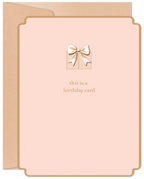 A modern blush pink greeting card with a detailed gold foil gift box and bow design on the front. Text reads 'this is a birthday card'. A kraft paper envelope peeks from behind.