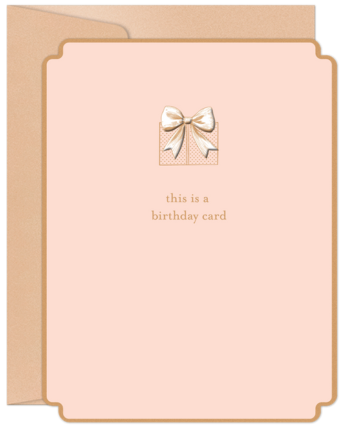 A modern blush pink greeting card with a detailed gold foil gift box and bow design on the front. Text reads 'this is a birthday card'. A kraft paper envelope peeks from behind.