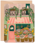 Front of an encouraging get well card featuring a charming, hand-drawn illustration of a Parisian florist shop with a pink awning, potted lemon trees, vibrant flowers, and a white cat peeking from an upstairs window. An 'OPEN' sign hangs on the door.
