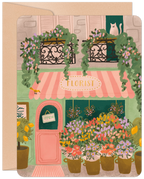 Front of an encouraging get well card featuring a charming, hand-drawn illustration of a Parisian florist shop with a pink awning, potted lemon trees, vibrant flowers, and a white cat peeking from an upstairs window. An 'OPEN' sign hangs on the door.