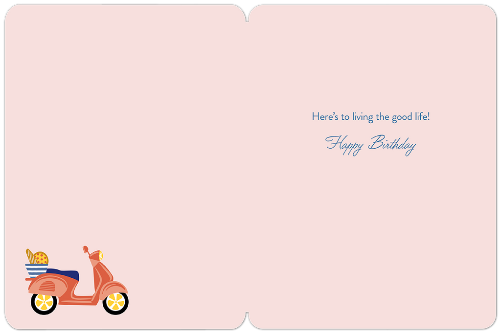 Inside of a light pink birthday card, showing the pre-printed message 'Here's to living the good life! Happy Birthday' in blue script, with a small scooter illustration in the bottom left corner.